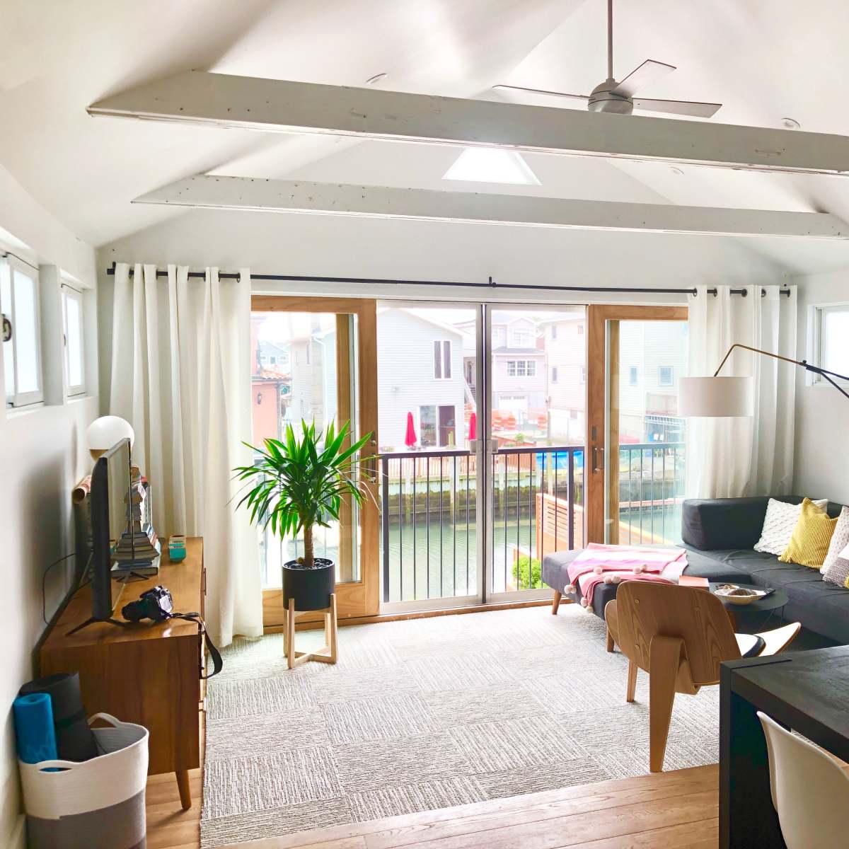 The living room features a sectional sofa, a dining table, and large windows overlooking a deck with a view of nearby houses.