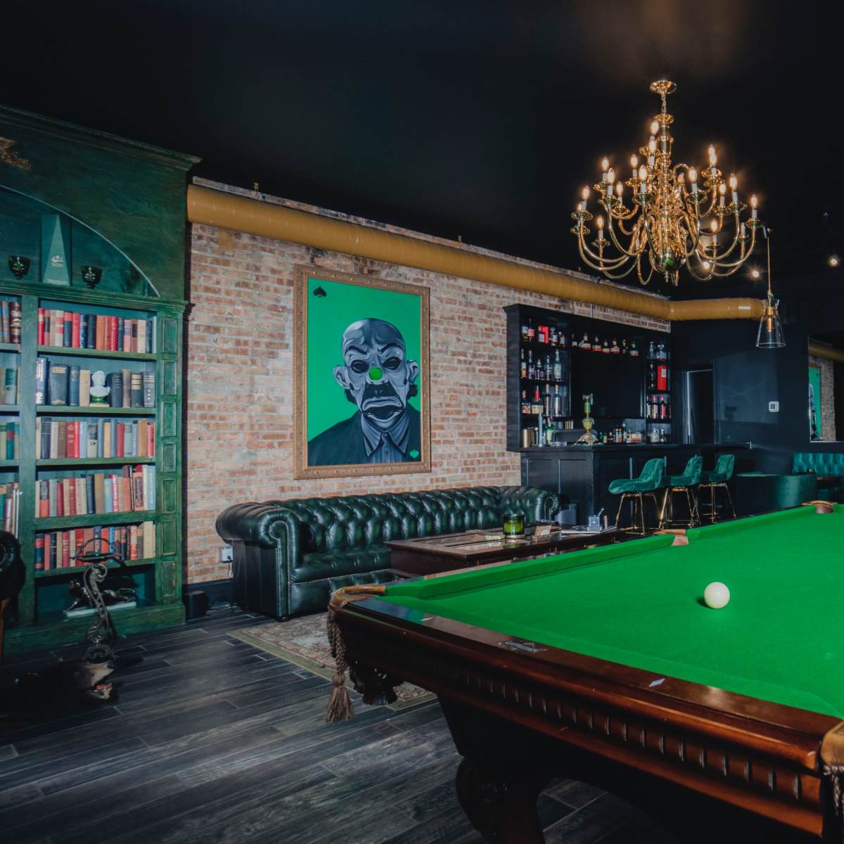A spacious game room features a green billiards table, a classic leather sofa, shelves lined with books, and a striking portrait on the wall.