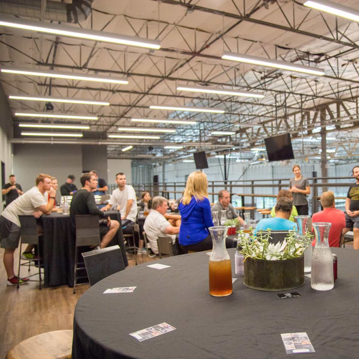 A group of people is seated and standing in a spacious indoor area with high ceilings, engaging in discussion while tables and drinks are arranged around them.