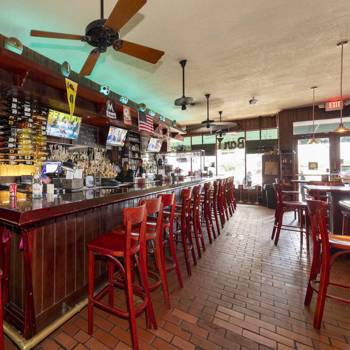 The interior of a bar with a wooden counter, several televisions displaying news, and red bar stools arranged along the counter.
