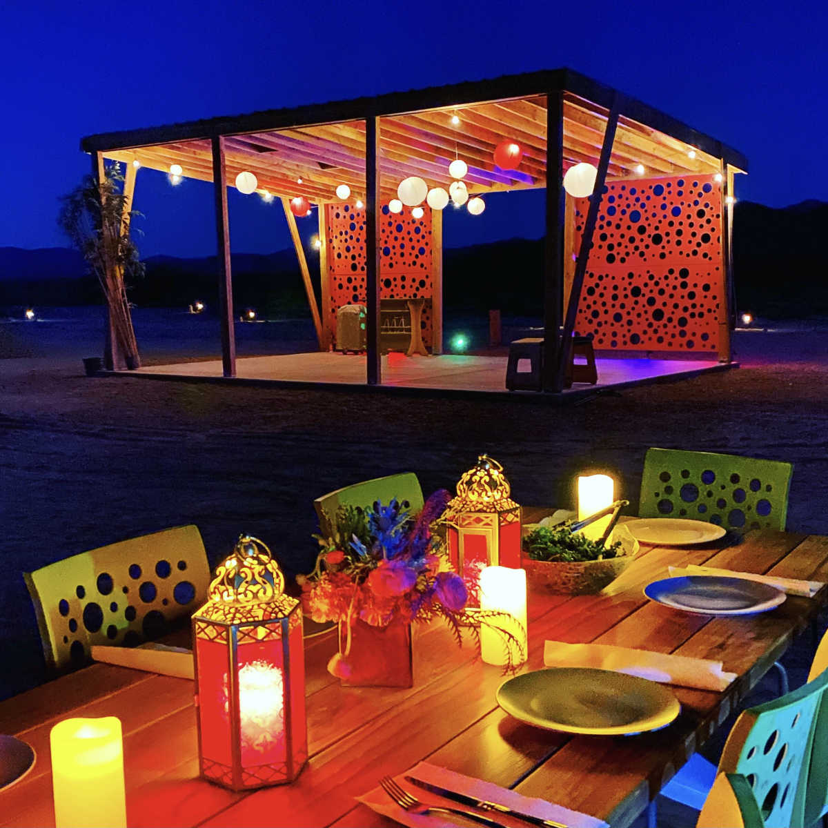 A brightly lit outdoor dining area features a long wooden table with lanterns and candles, set against a decorated pavilion under a night sky.