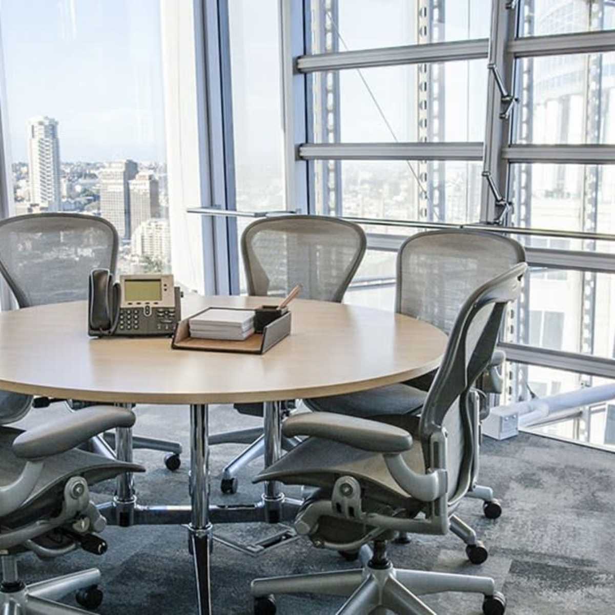 A modern conference room features a round table surrounded by ergonomic chairs with large windows offering a city view.
