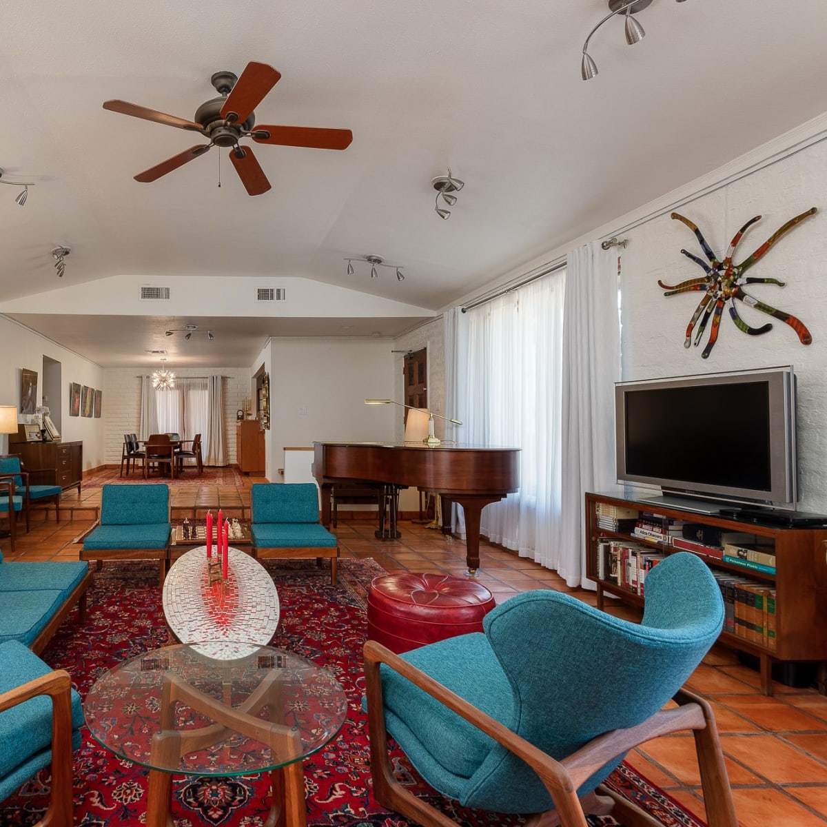 A spacious living area features a circular arrangement of teal armchairs around a glass table, with a piano and TV in the background.