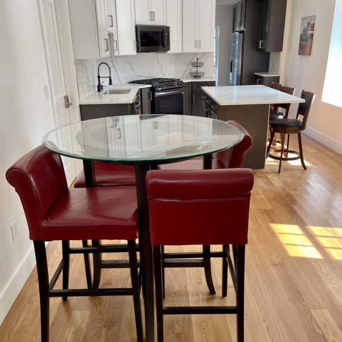 A modern kitchen with a glass-top dining table surrounded by red high-back chairs, along with black and white cabinetry in the background.