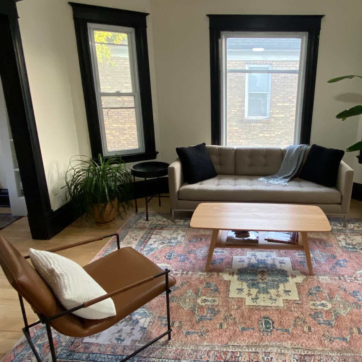 A living room with a tan couch, a wooden coffee table, a patterned area rug, and large windows, accompanied by a potted plant and a chair.