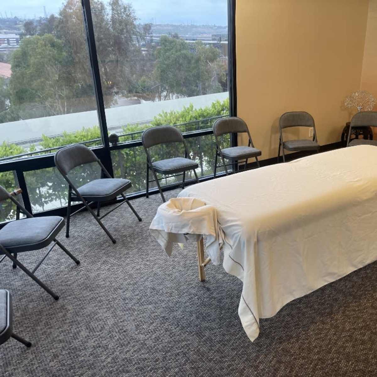 A massage table covered with a white cloth sits in the center of a room surrounded by eight gray chairs.