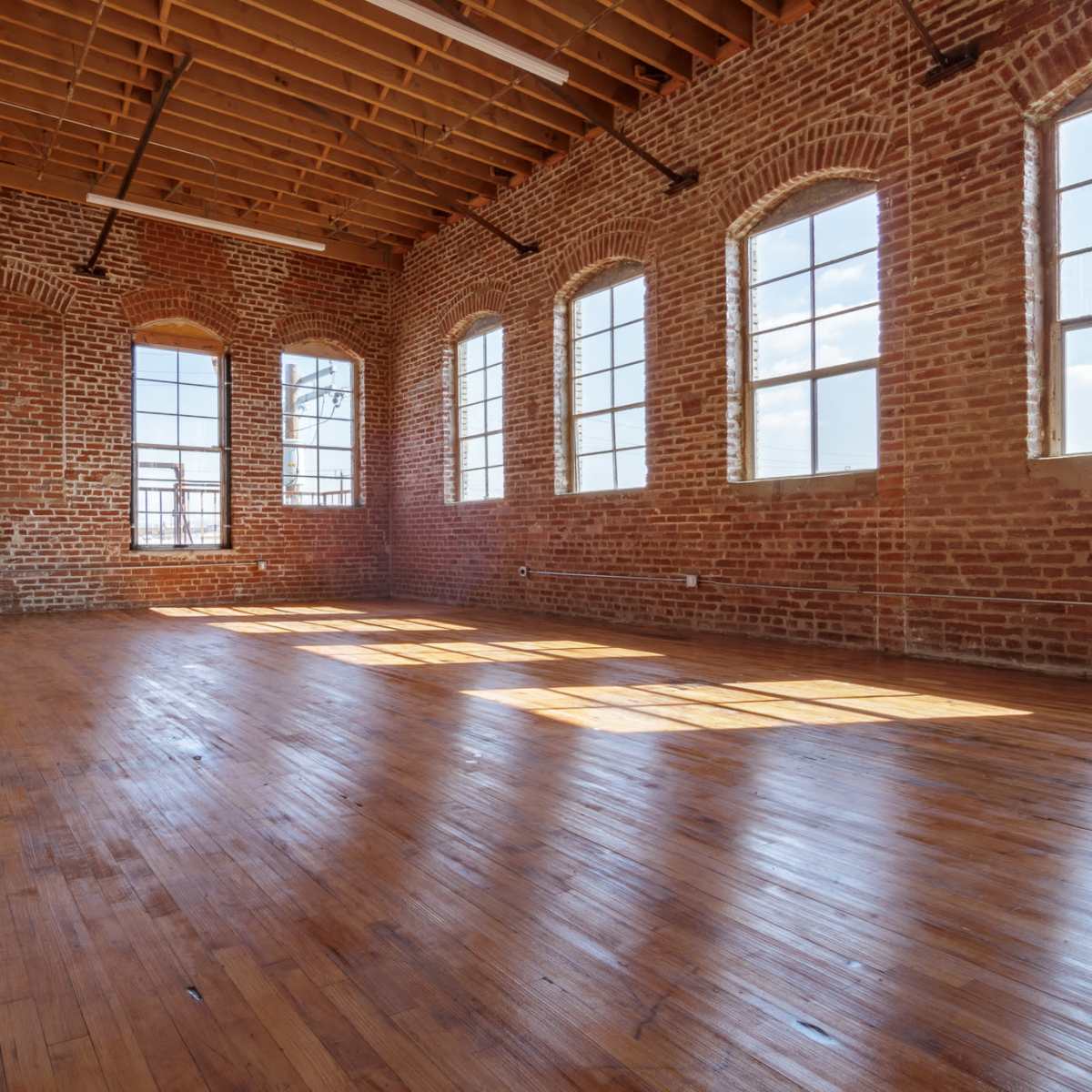 An empty room with exposed brick walls, wooden floors, and large windows allowing natural light to fill the space.