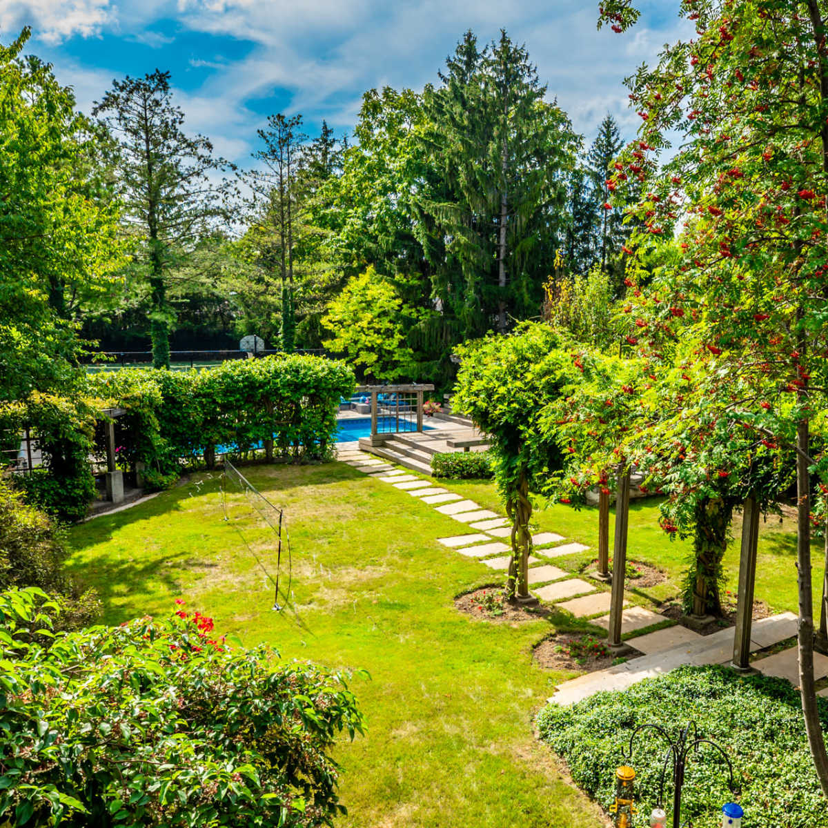 A landscaped garden featuring a stone pathway leading to a swimming pool, surrounded by greenery and trees.