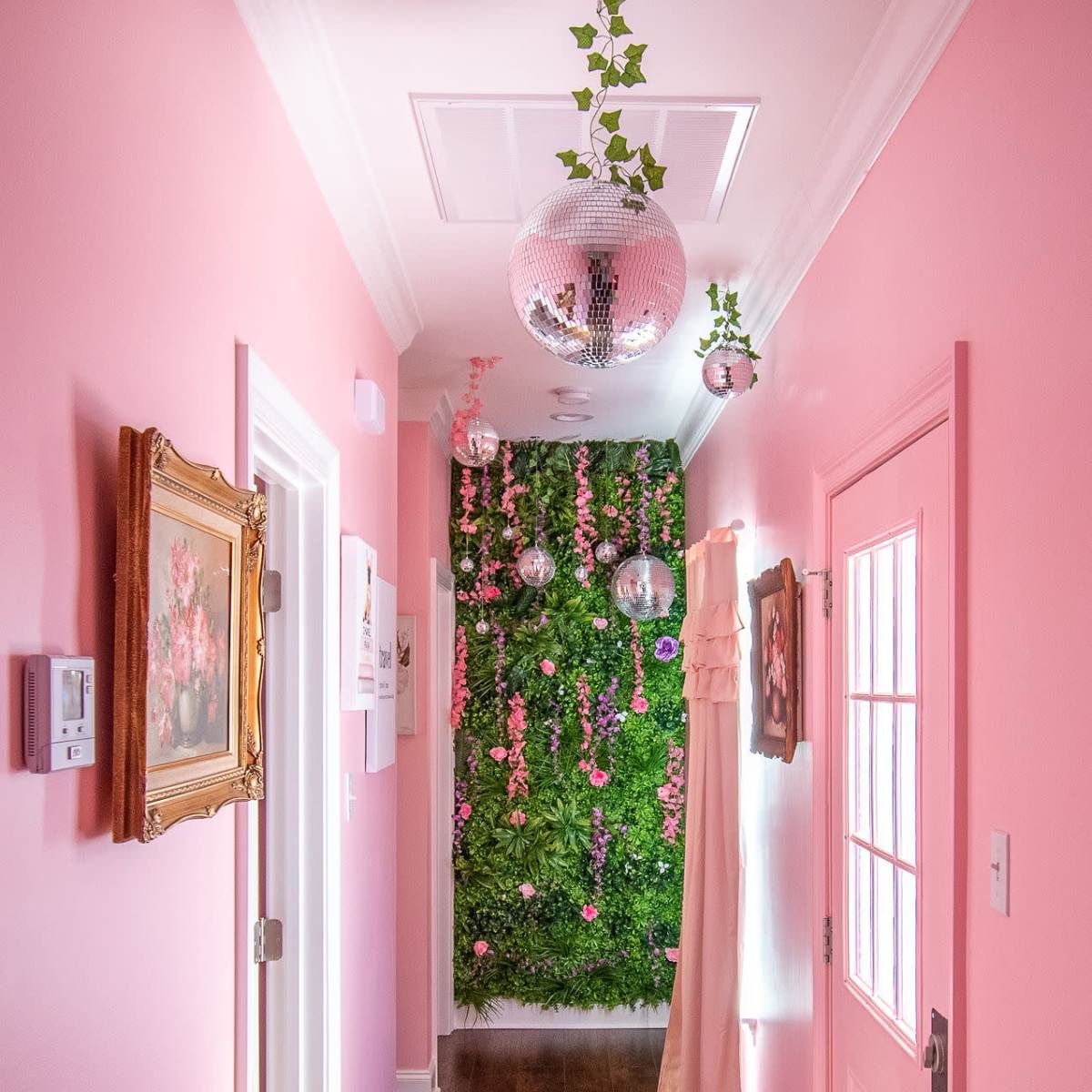 A pink hallway features a green vertical garden along one wall, adorned with hanging plants and disco balls, leading to a brightly colored door.