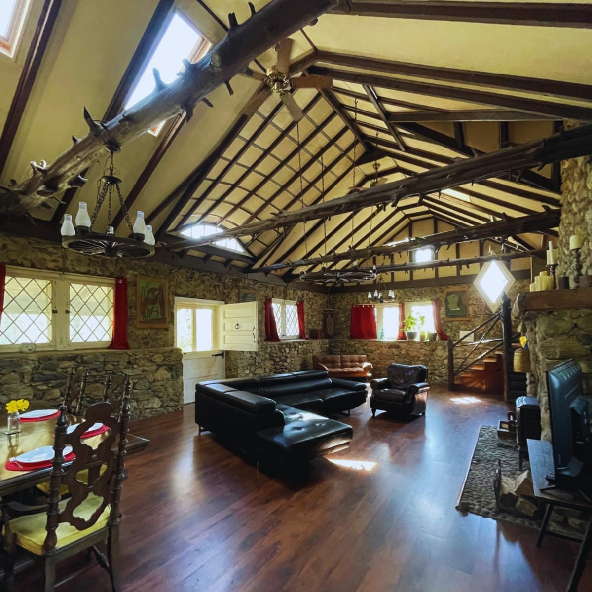A spacious interior of a rustic living area with a wooden ceiling, stone walls, a black leather sofa, and a dining table set for a meal.