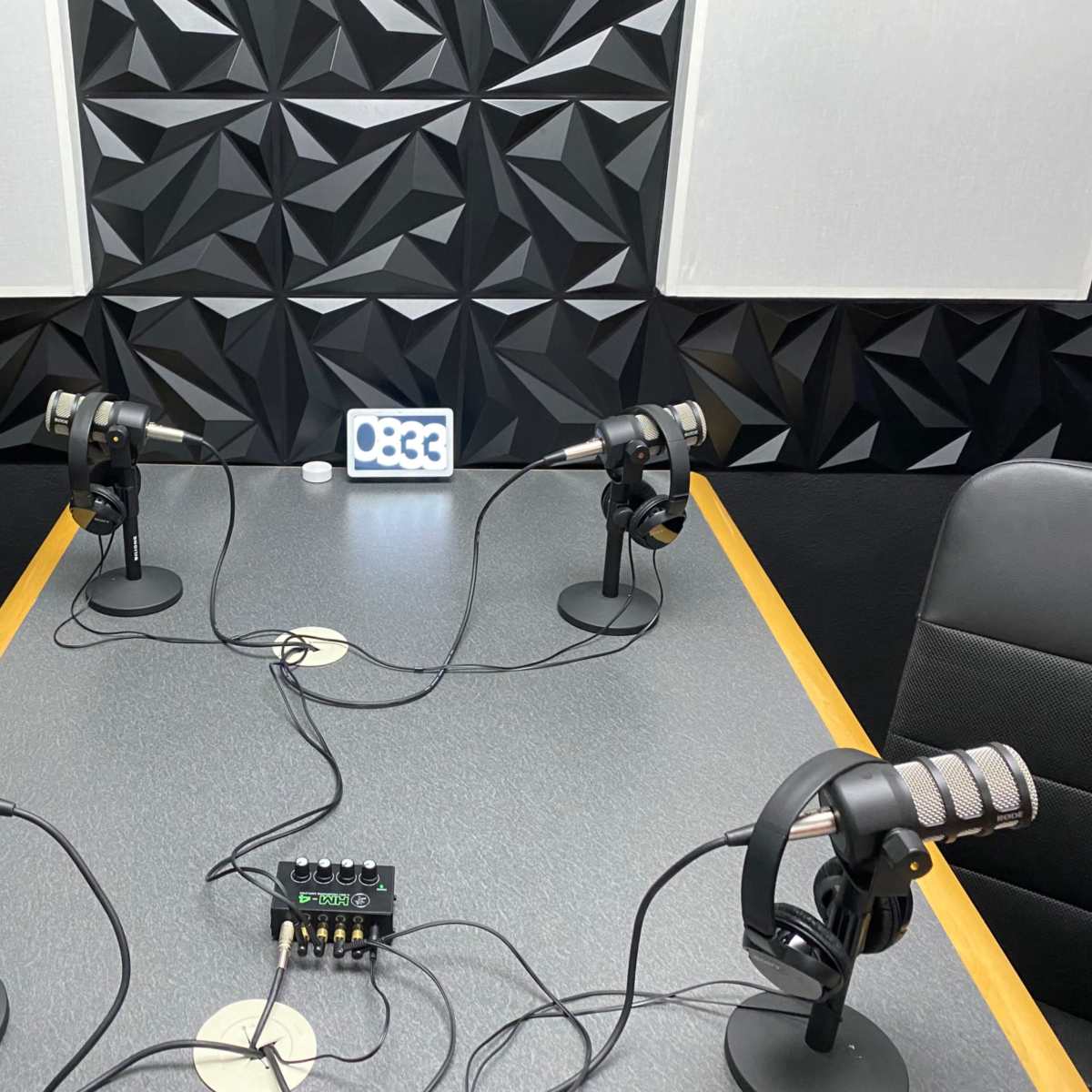 A podcast recording studio with four microphones, headphones, and a mixer on a table surrounded by textured black walls.
