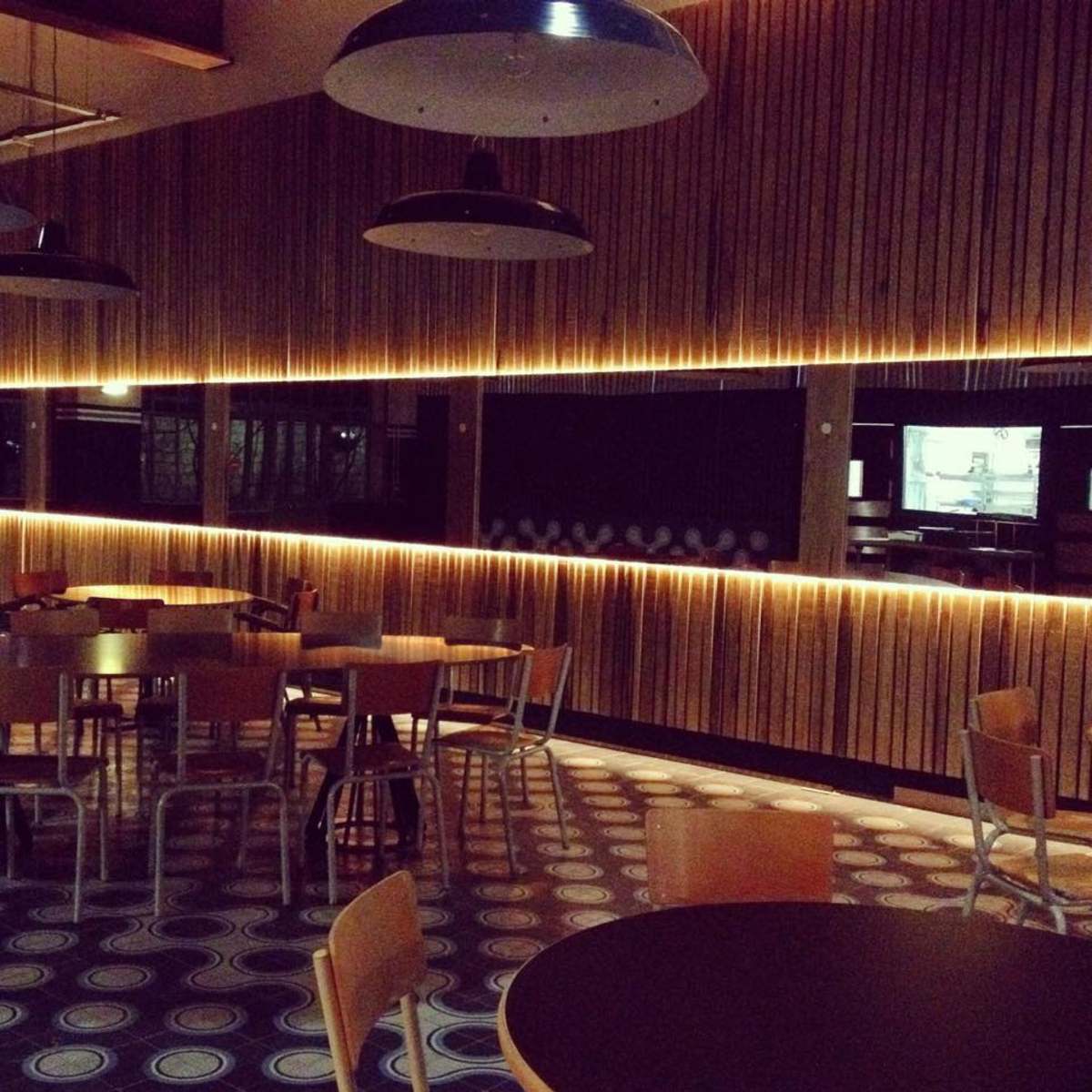 A modern cafe interior features wooden paneling, circular patterned flooring, and empty tables illuminated by warm overhead lighting.