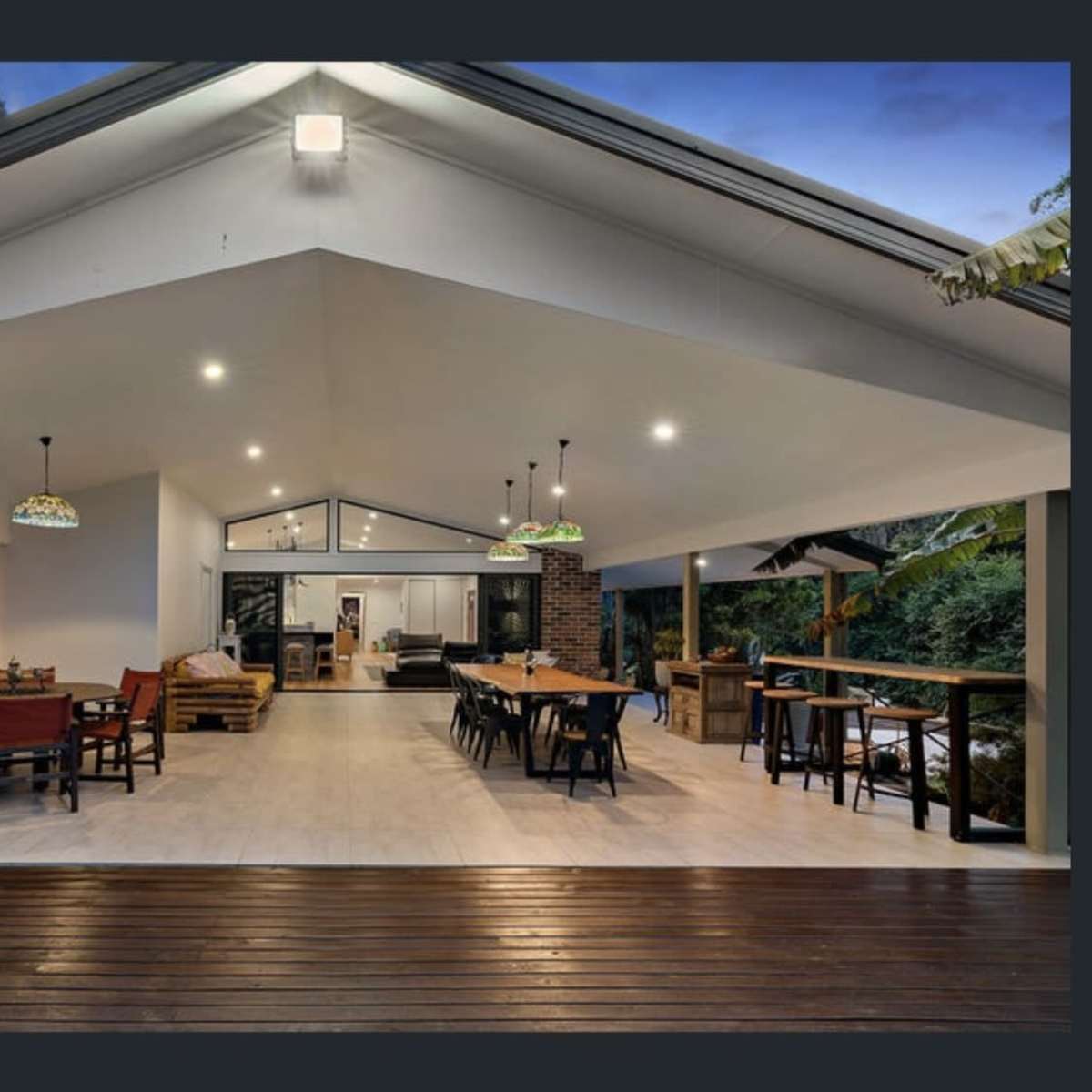 A spacious outdoor living area with wooden flooring, featuring dining tables and chairs under pendant lights, connected to a modern interior space.