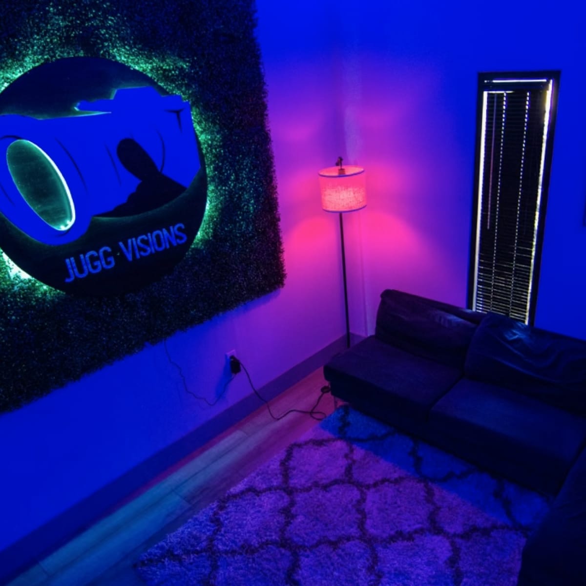 A low-lit room with a green neon sign reading "Jugg Visions," a sofa, a lamp casting colorful light, and a rug on the floor.