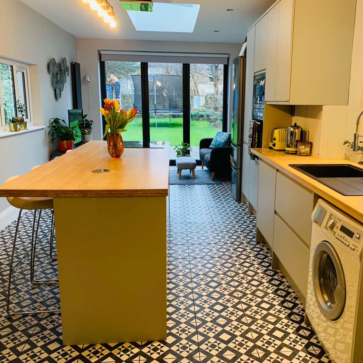 A modern kitchen with a central island, equipped with a sink and a washing machine, leading to a garden view through large glass doors.
