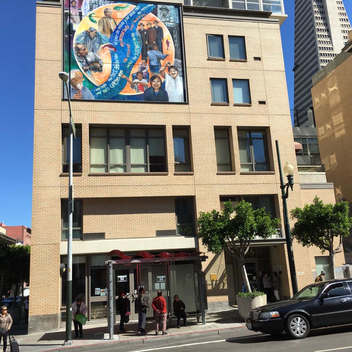 A building with a large mural on its exterior stands at a street corner, with people waiting at a bus stop in front.