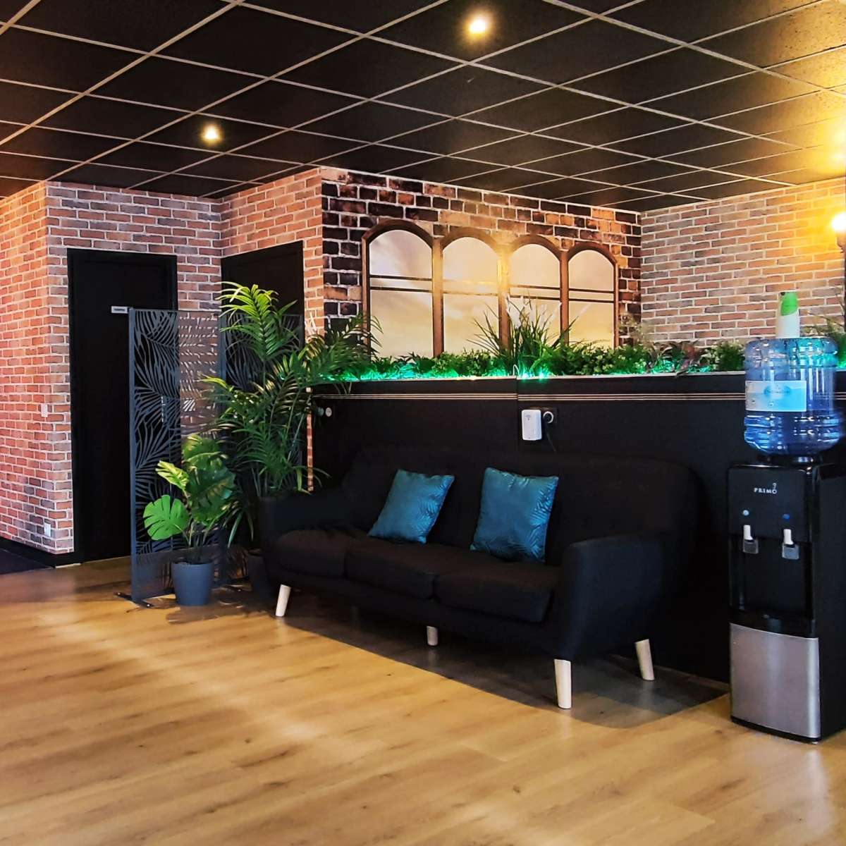 A modern office lobby with a reception area, seating, brick walls, and plants, featuring a water cooler and counter.