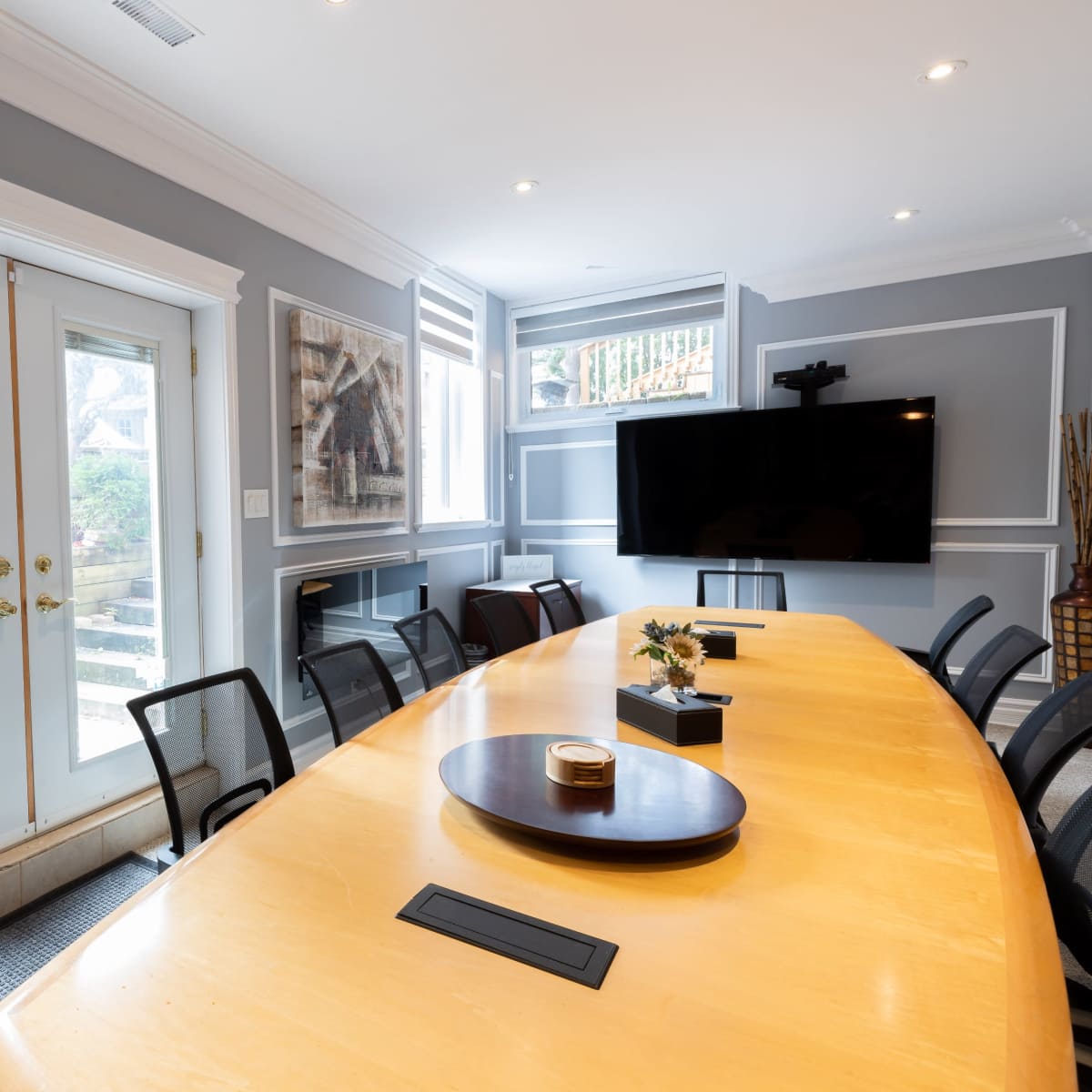A large conference room features a long wooden table surrounded by black chairs, with double doors leading to an outdoor area and a mounted television on the wall.