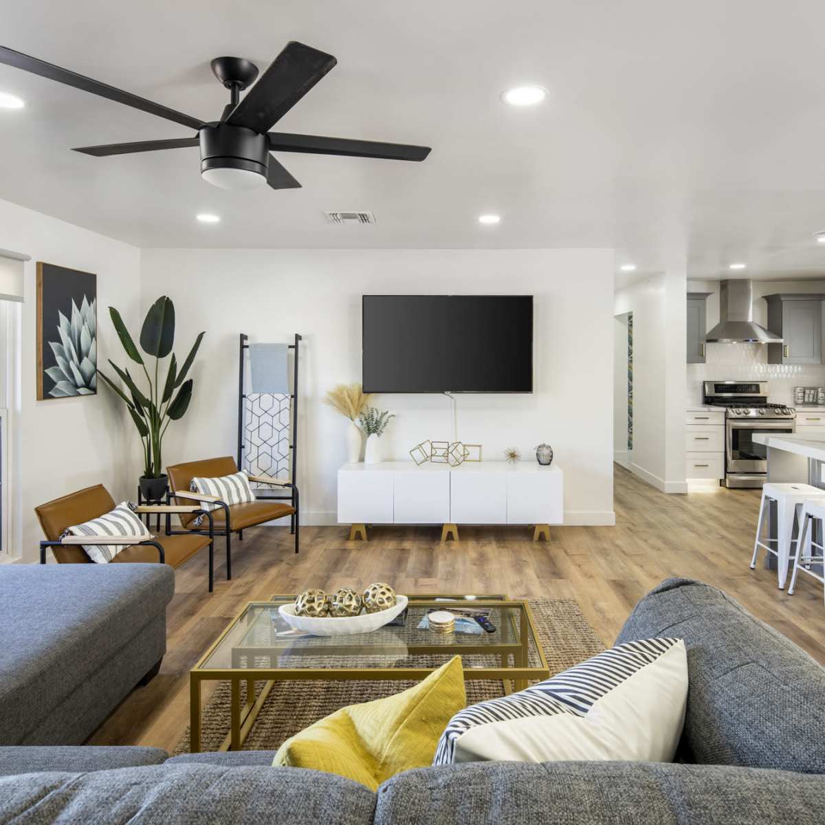 A modern living room with a gray sectional sofa, a large wall-mounted television, and an open kitchen area featuring white cabinetry and bar stools.