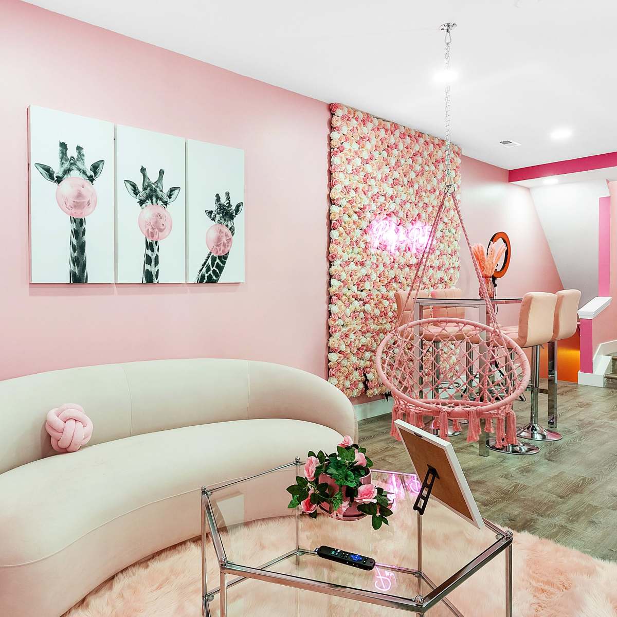 A stylish pink room featuring a curved sofa, floral artwork, a hanging chair, and a glass coffee table with a plant.