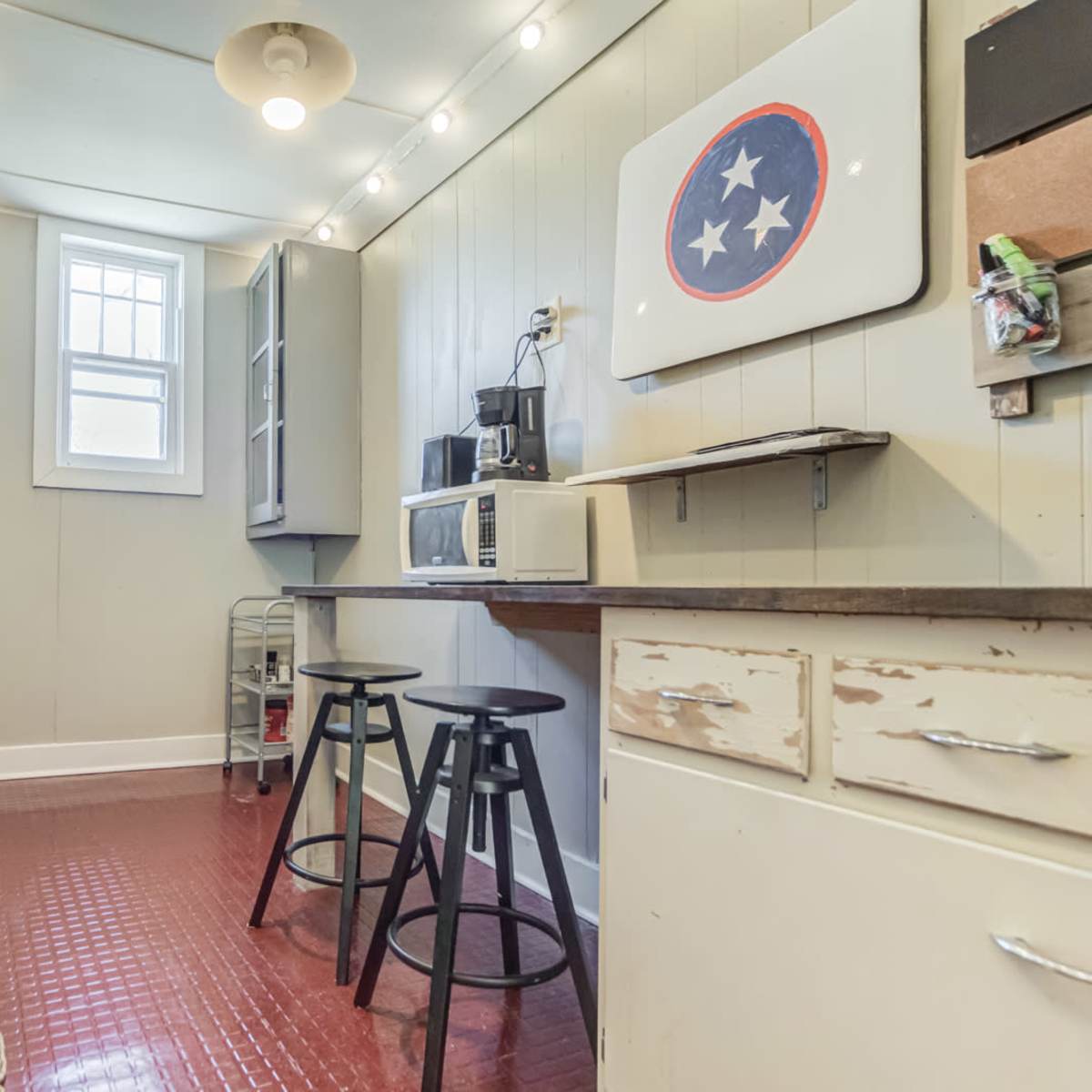 A compact kitchen with a black stove, microwave, and two black stools next to a countertop, featuring light-colored walls and checkered curtains.