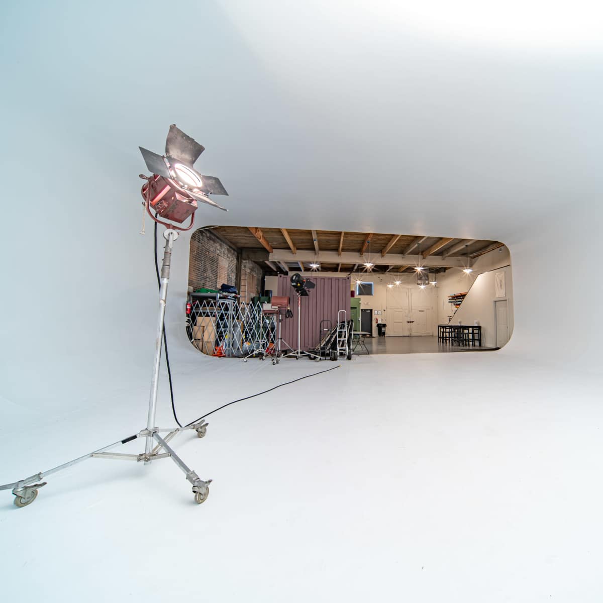 A spacious, white studio with a lighting fixture on a stand in the foreground and various equipment visible in the background.