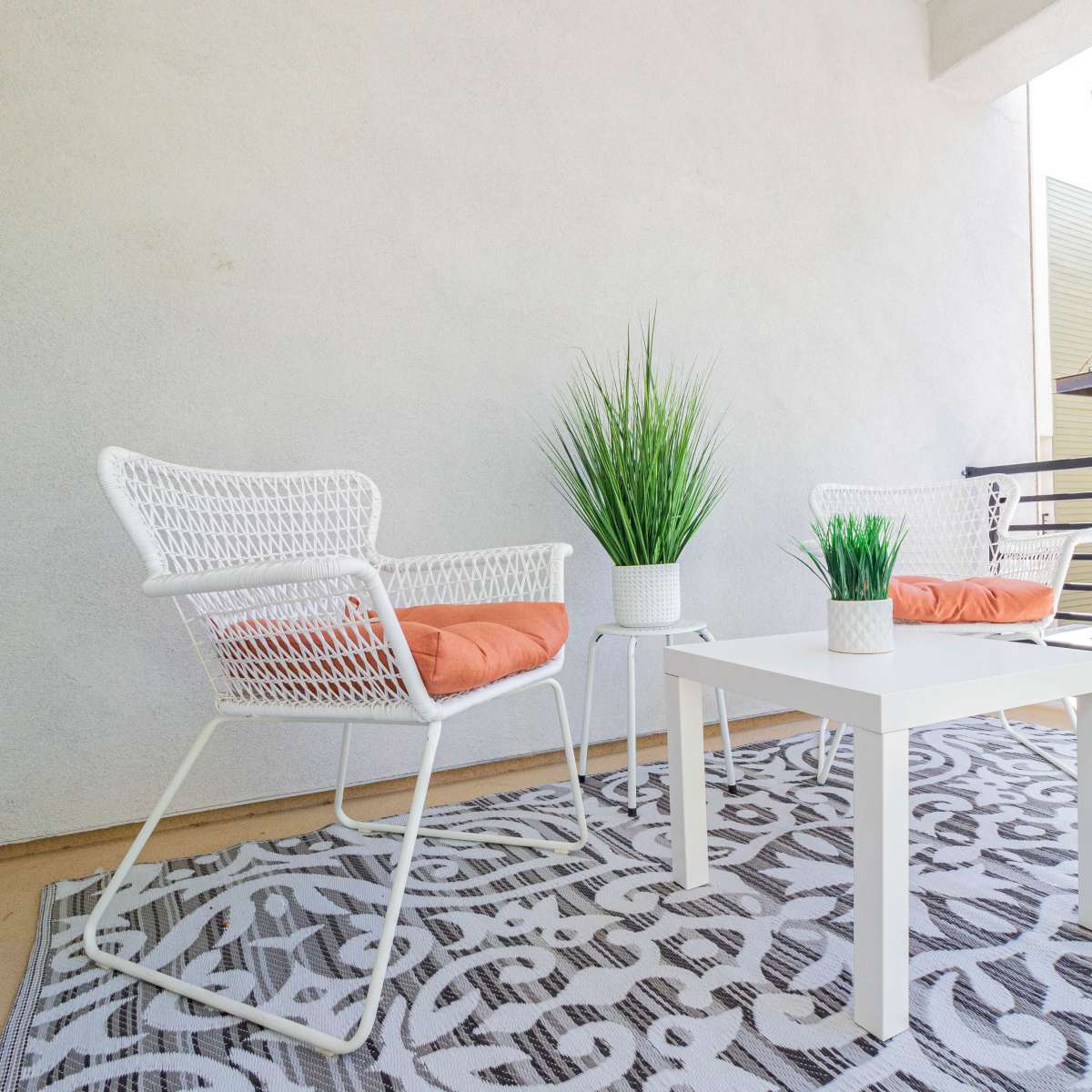 A balcony with two white wicker chairs, an orange-cushioned table, and potted plants arranged on a patterned rug.