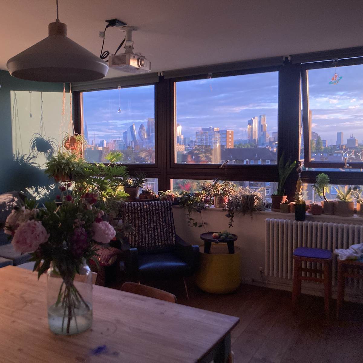 A cozy living room features a wooden table, a vase of flowers, and large windows showcasing a city skyline at sunset.