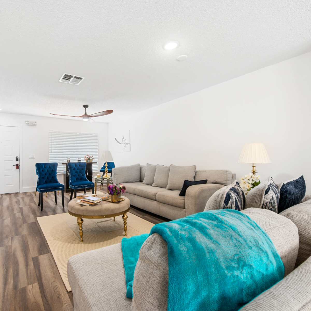 A bright living room features a light-colored sofa, a round coffee table with flowers, two blue armchairs, and a ceiling fan.
