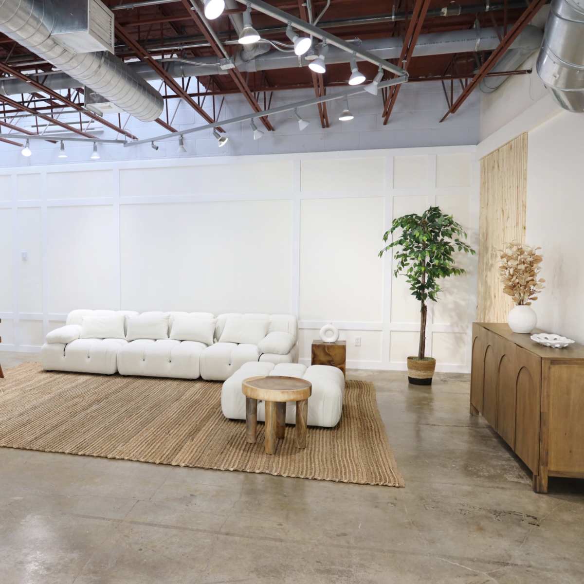 A spacious, minimalist interior features a white sectional sofa, two wooden chairs, a round wooden table, and a large area rug on polished concrete flooring.