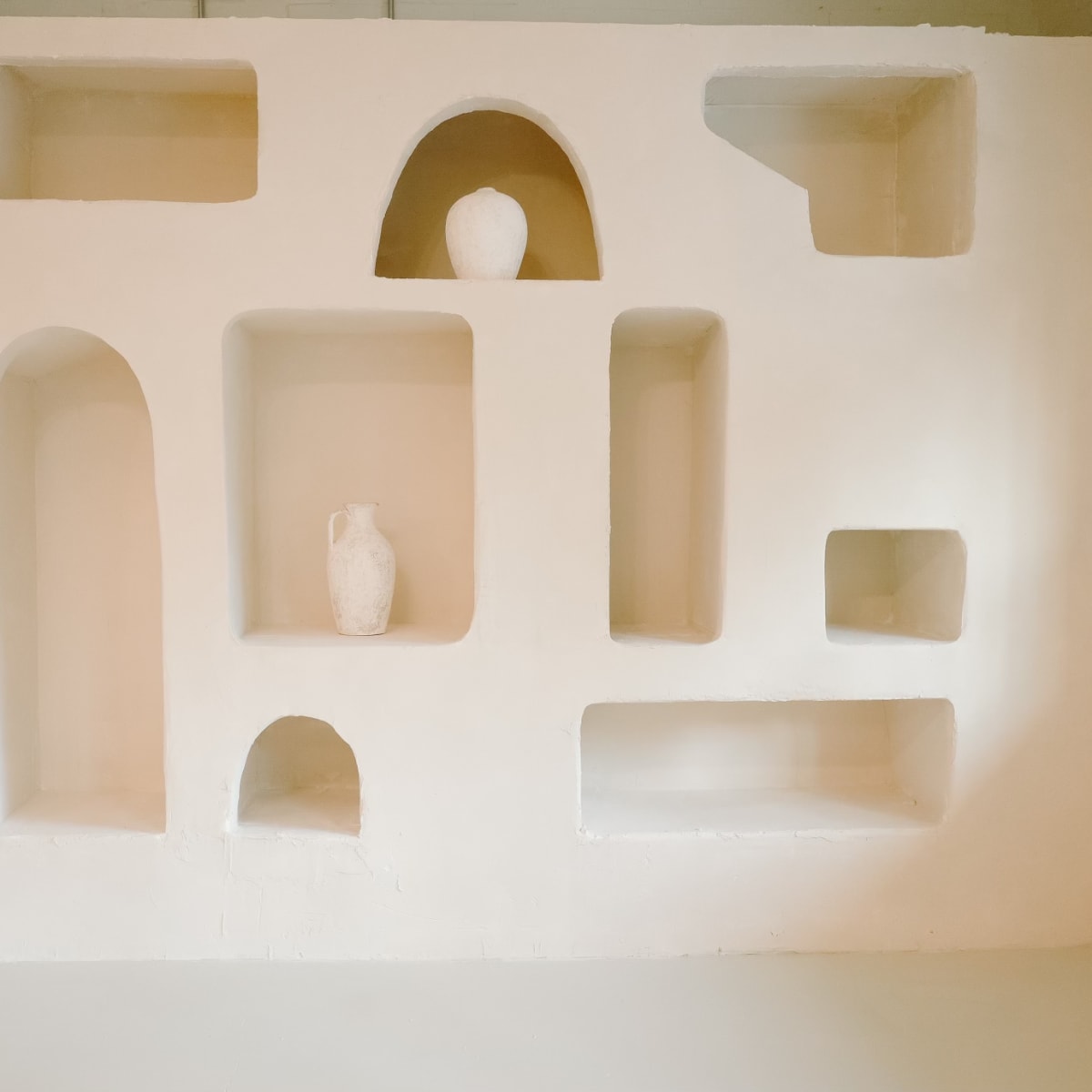 A minimalist wall display features various recessed niches holding white ceramic vases and shapes.