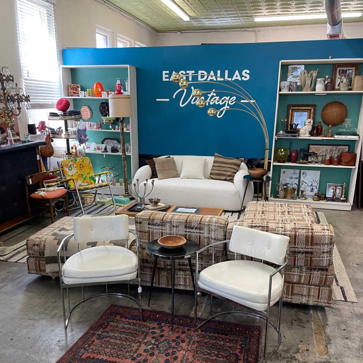 A vintage furniture store display featuring a sofa, patterned ottomans, and white chairs, set against a bright blue wall with shelves of decor items.