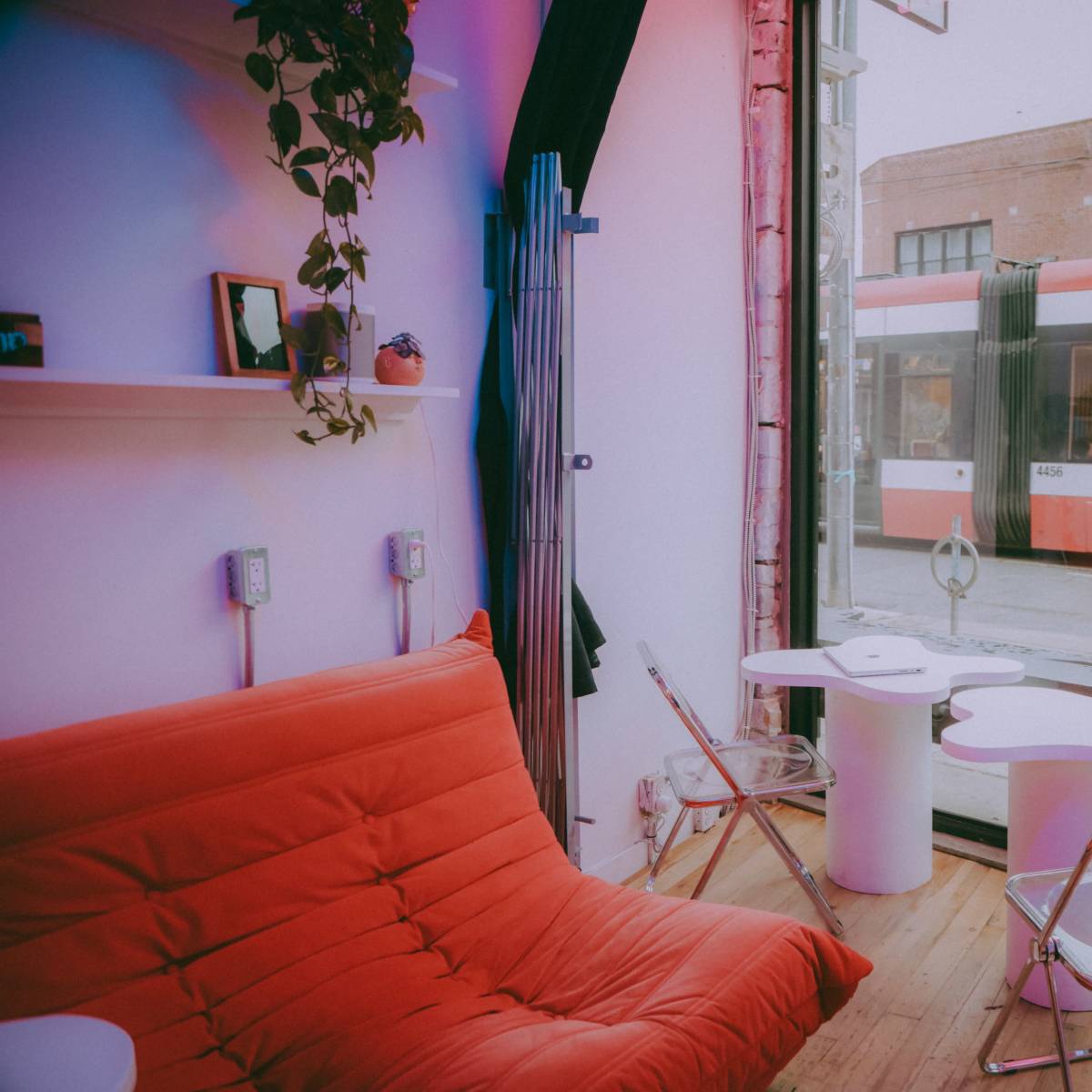 A red couch and modern chairs are arranged in a bright, minimalist interior next to a large window with a view of a tram passing by.