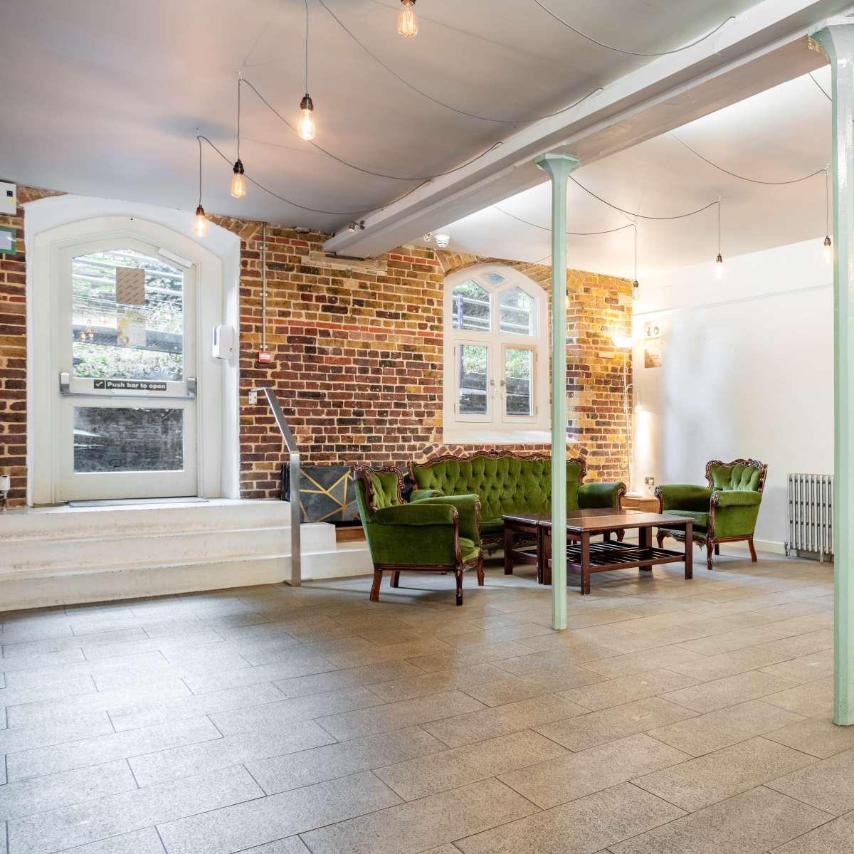 An interior space with a green seating area, a wooden table, and a doorway leading to another room, all against a backdrop of exposed brick walls and hanging light bulbs.