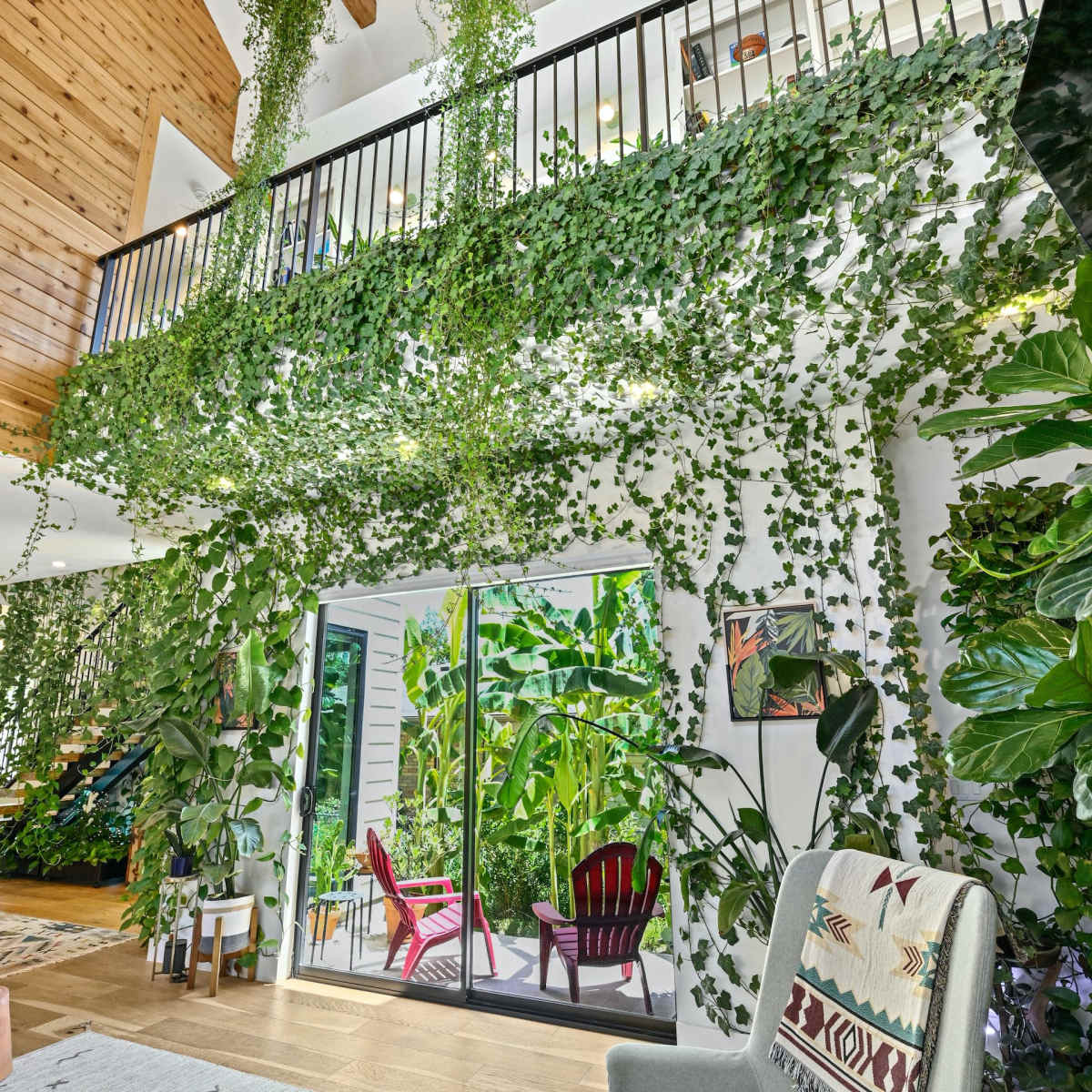 The interior of a brightly lit, two-story living room features tall plants cascading from the balcony and large sliding glass doors leading to a garden patio.