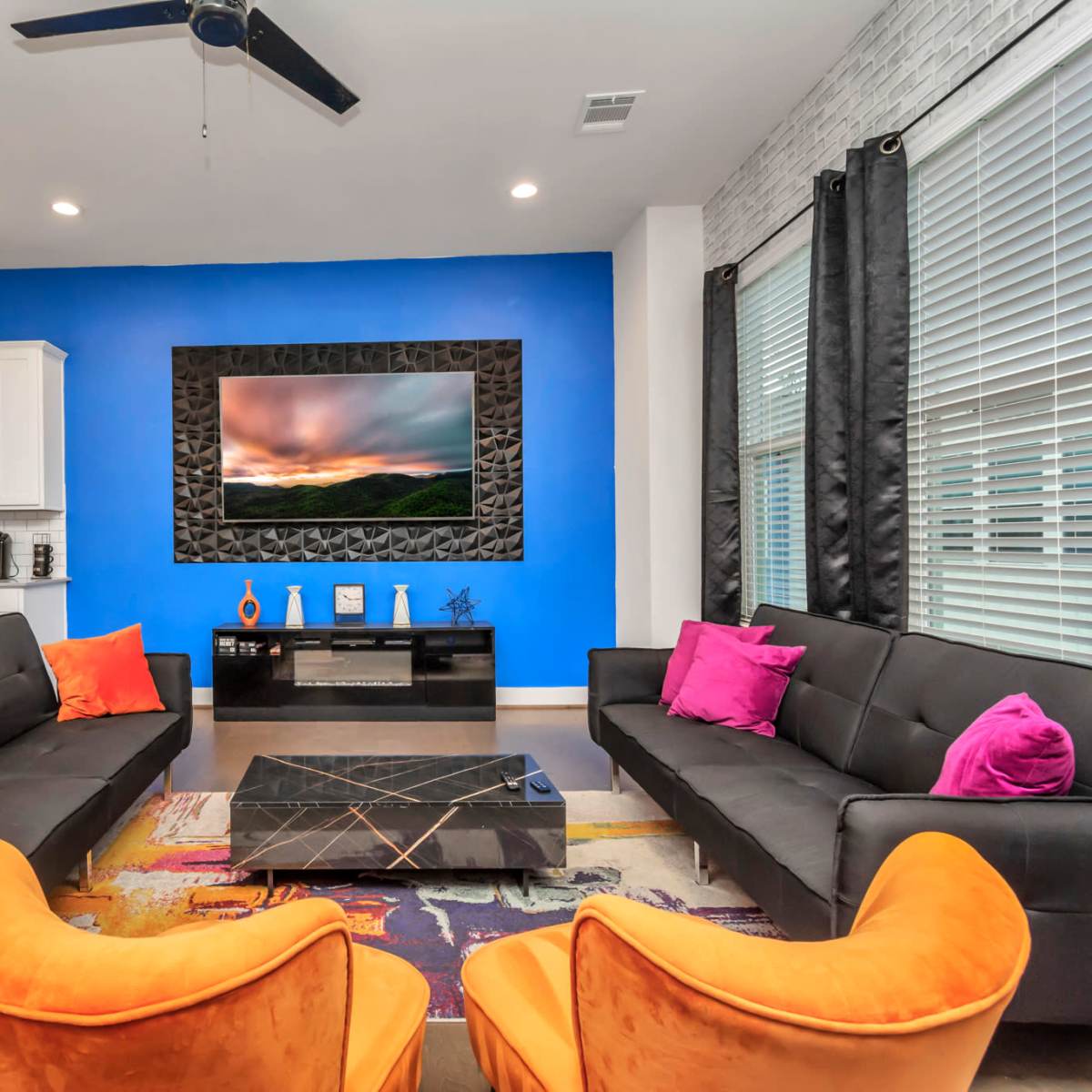 A modern living room features a blue accent wall, two black sofas with colorful cushions, an orange armchair, a coffee table, and a mounted television above a sleek fireplace.