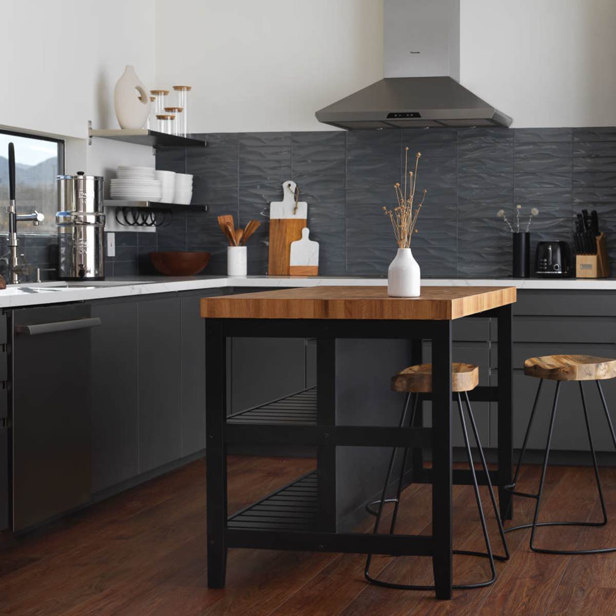 The modern kitchen features a central wooden island with two metal stools, gray cabinetry, stainless steel appliances, and a textured backsplash.