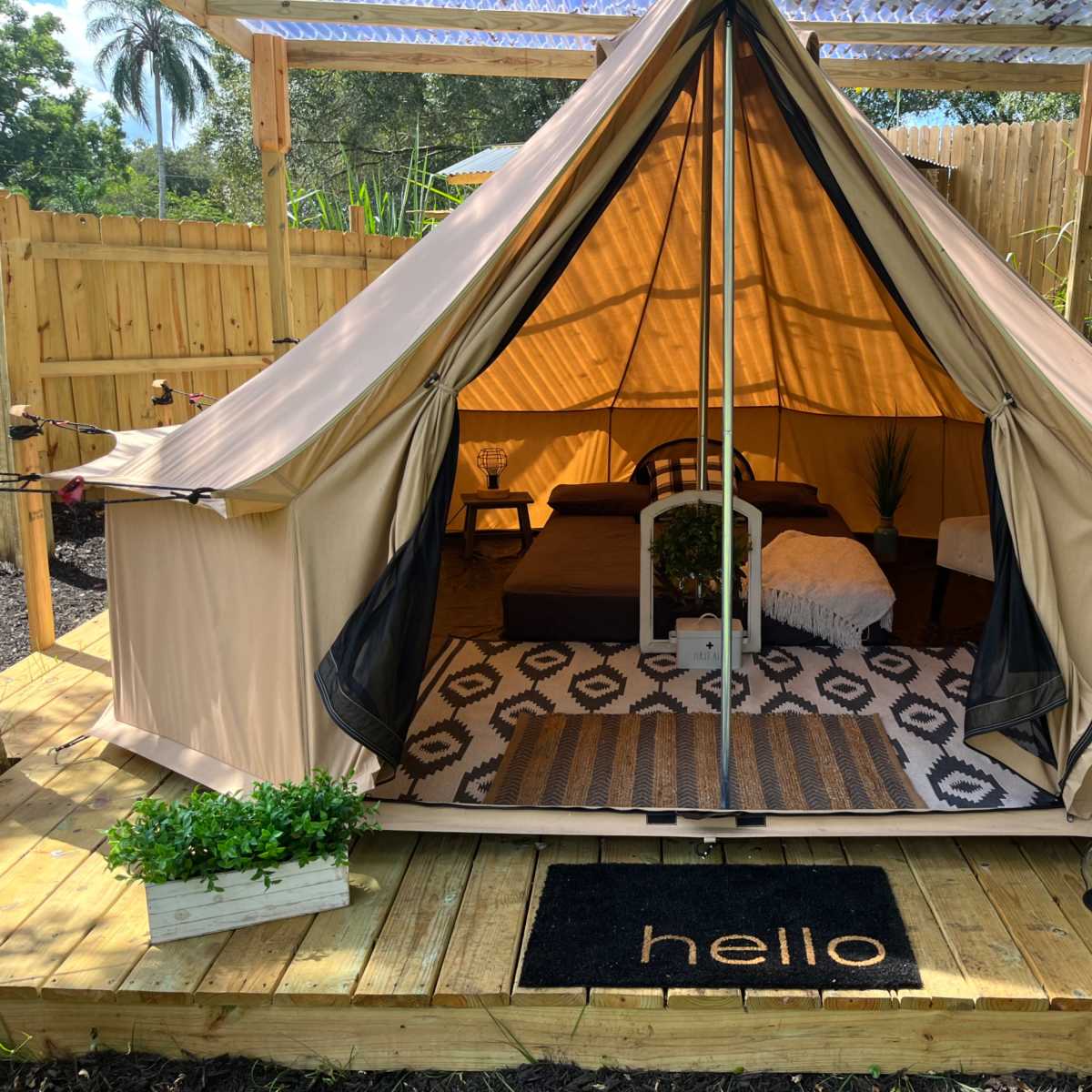A beige tent set up on a wooden deck, featuring a bed, a decorative rug, a potted plant, and a welcome mat that reads "hello."