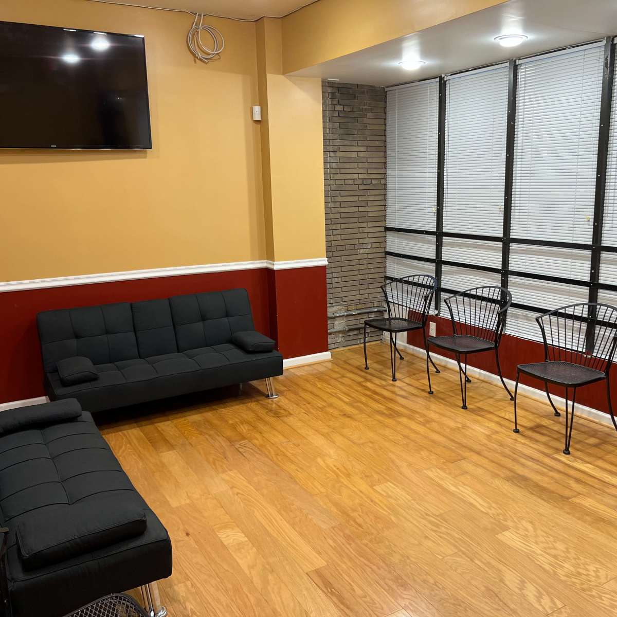 The room features a black futon, a television mounted on the wall, and several metal chairs arranged along a windowed wall.