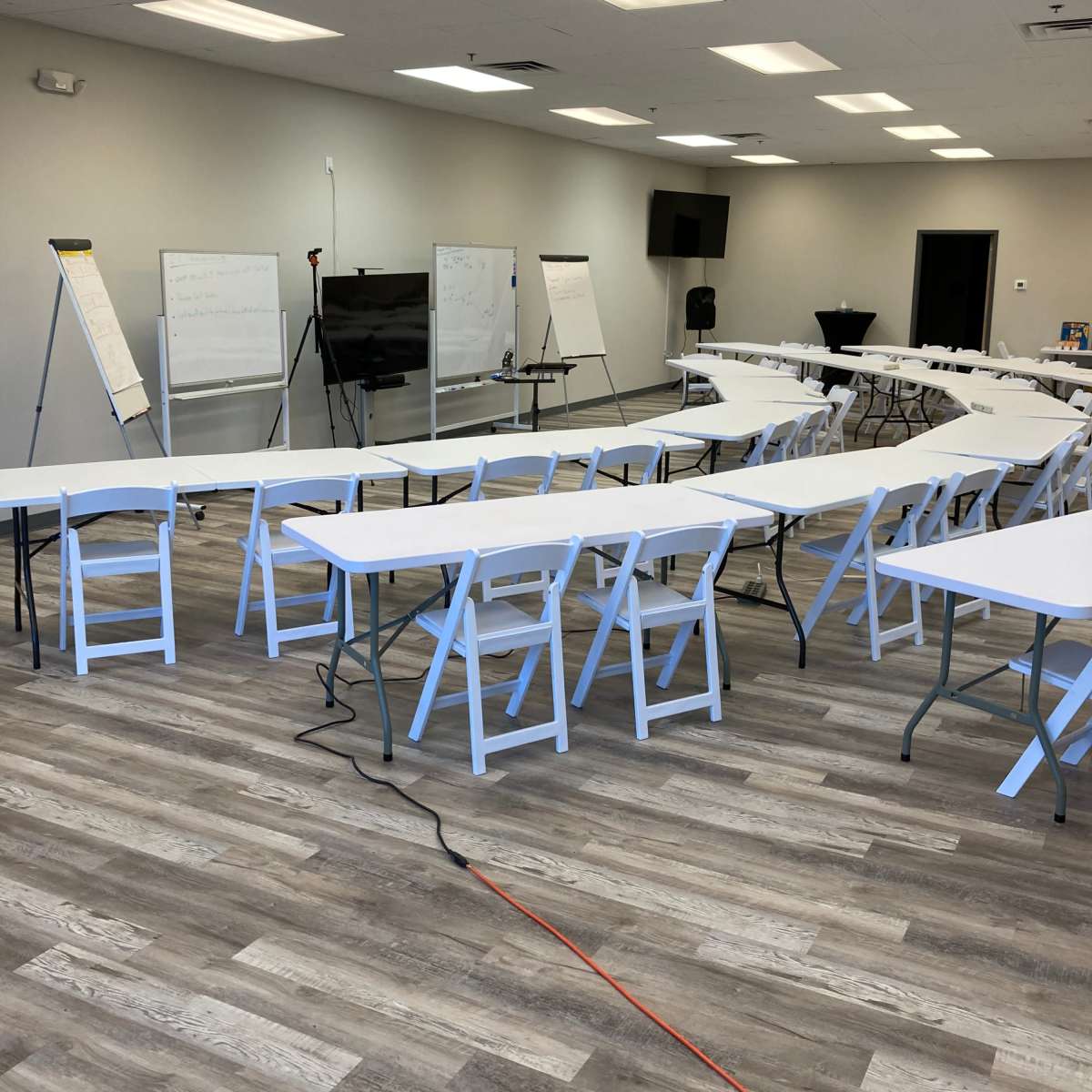 A spacious training room contains several white tables arranged in rows, along with flip charts and a presentation screen at the front.