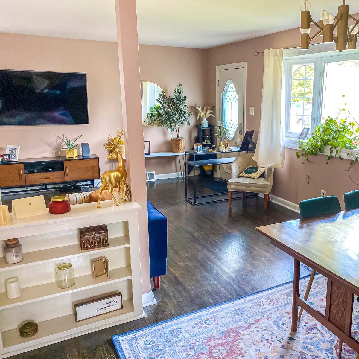 A well-decorated living space featuring a dining area with a wooden table and chairs, a television mounted on the wall, and various plants and decor items throughout.