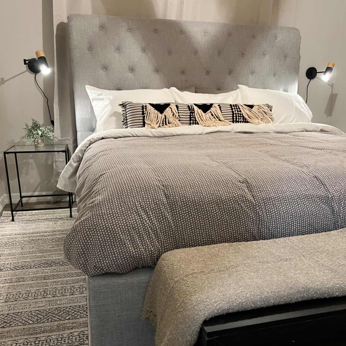 A neatly made bed with a gray upholstered headboard, a patterned blanket, and decorative pillows, accompanied by a small nightstand and a bench at the foot of the bed.
