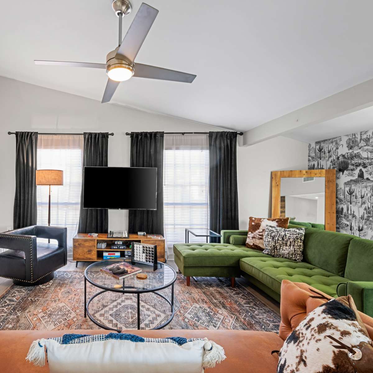 A modern living room features a green sectional sofa, an orange couch, a circular coffee table, and a mounted television above a fireplace, with large windows dressed in black curtains.