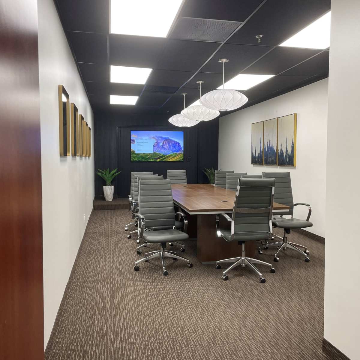 A modern conference room with a large wooden table, six gray leather chairs, a mounted screen displaying a landscape, and artwork on the walls.