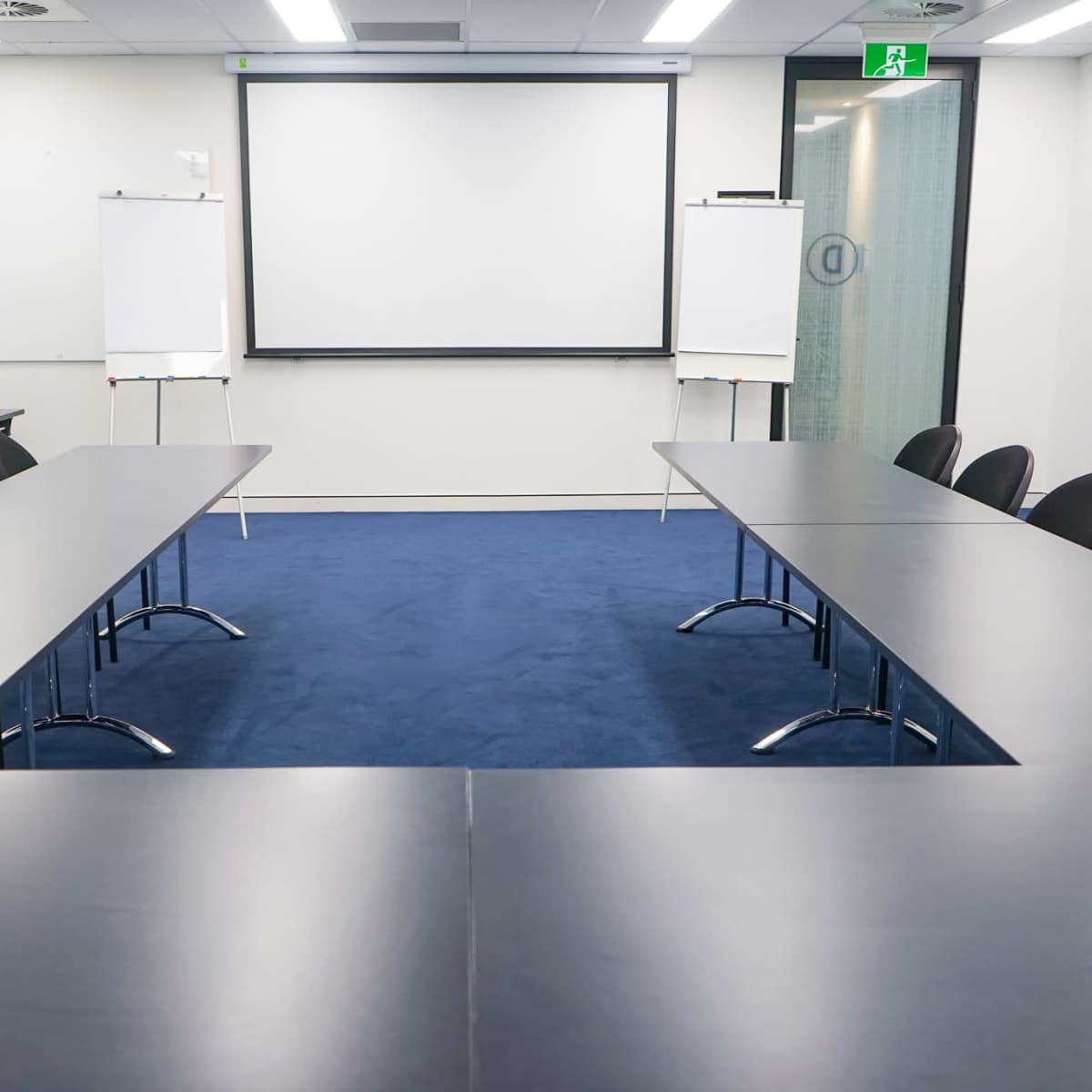 A conference room is set up with two rectangular tables facing a presentation screen, flanked by empty chairs and whiteboards.