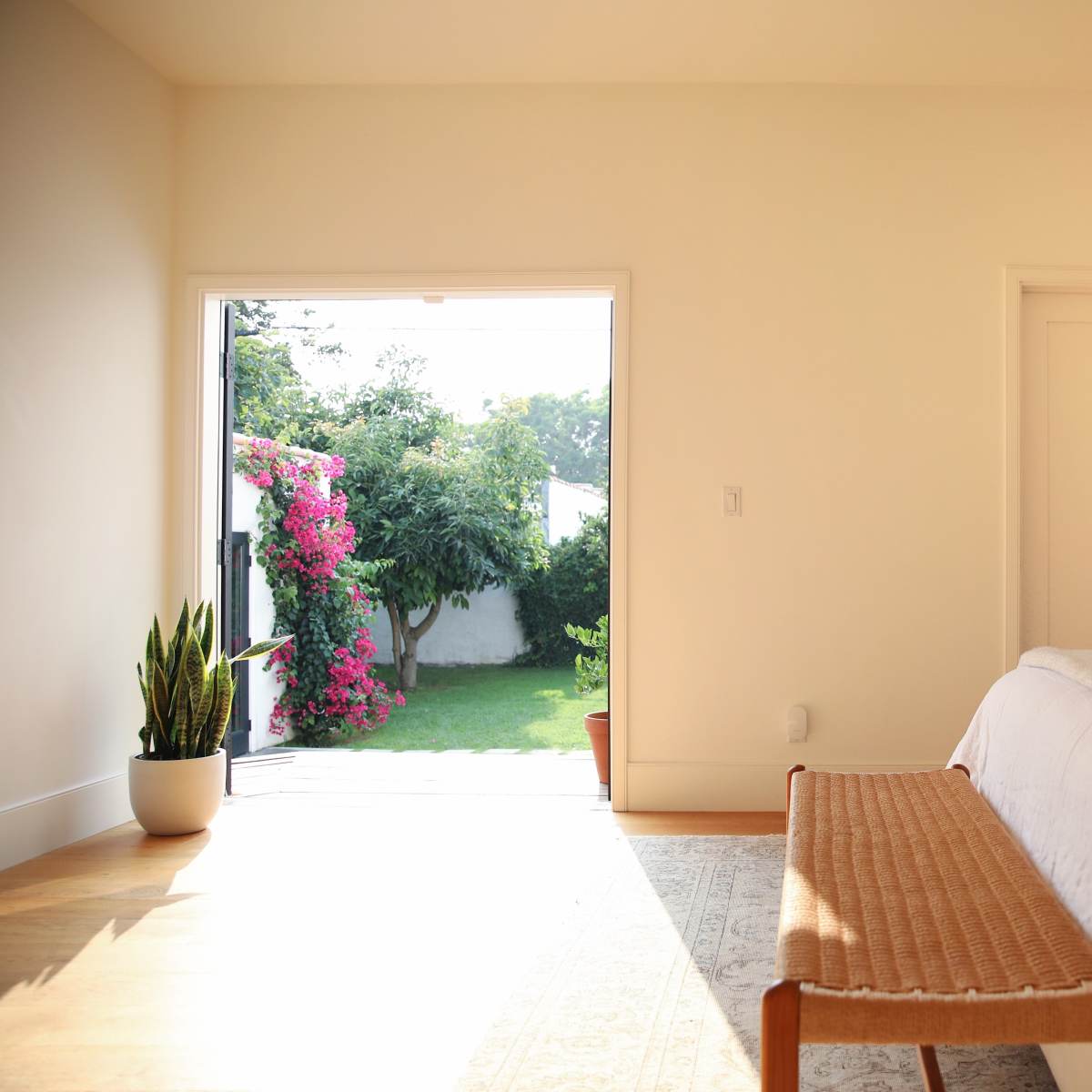 A bright bedroom features a bed with a light-colored coverlet, a wooden bench at its foot, and a door that opens to a garden with blooming plants.