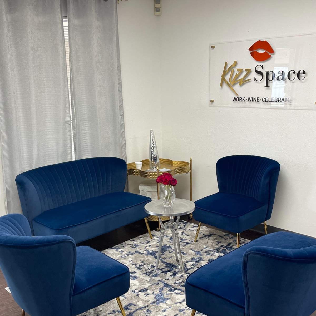 A small, modern seating area featuring a blue velvet sofa and chairs arranged around a glass coffee table, placed on a patterned rug against a white wall with a decorative sign.