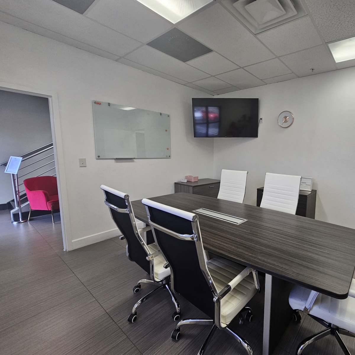 A modern conference room with a long wooden table, six white ergonomic chairs, a wall-mounted television, and a whiteboard.