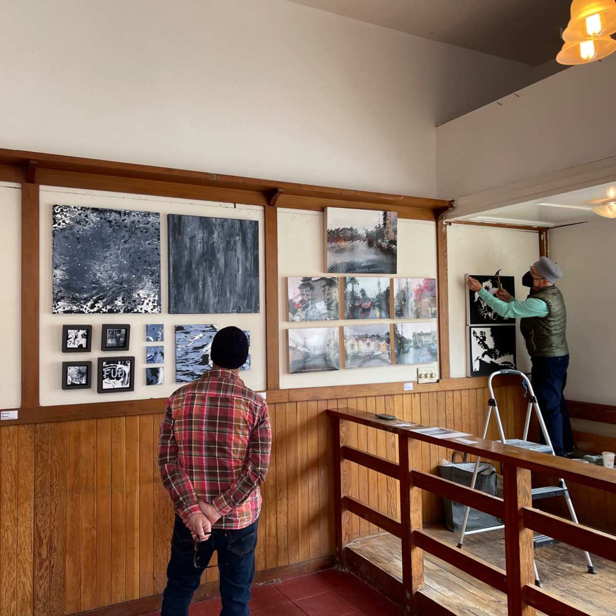 Two people are inside an art gallery; one man stands observing the artwork on the wall while another man is hanging a painting.