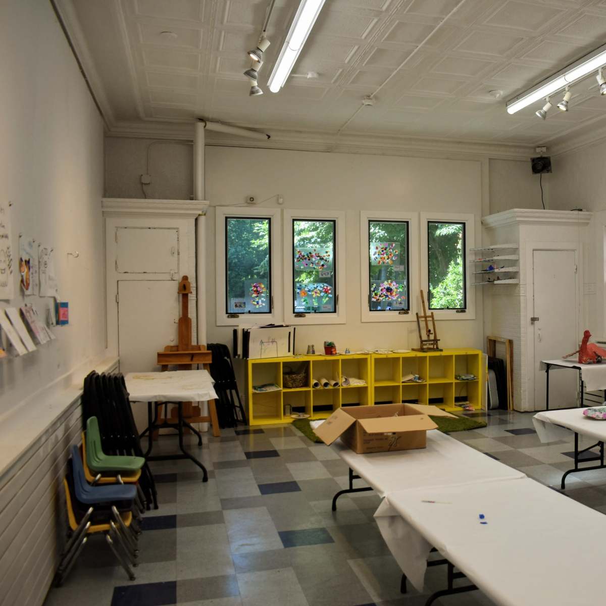 A spacious art classroom with tables covered in white cloths, colorful stained glass windows, and yellow storage shelves along the wall.